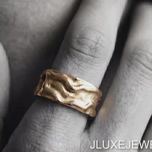 May include: A wide gold band ring featuring an irregular, wavy, and textured surface. The ring is worn on a finger, and the text JLUXEJEWELS is visible in the lower right corner, with the brand logo in the upper left corner of the image.