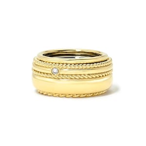 May include: A gold ring featuring a multi-band design with a smooth central band, a rope-textured band, a beaded band, and a single small round clear gemstone set into the metal. The ring has a polished finish and a layered, stacked appearance.