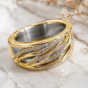 May include: A wide band ring featuring a two-tone design with a polished silver-colored inner band and a gold-colored outer layer. The gold exterior is composed of multiple overlapping, curved, and textured ribbons encrusted with small clear stones.