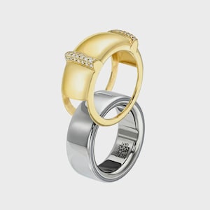 Könnte beinhalten: Ein goldener Ring mit einer glatten, gewölbten Oberseite und zwei Bändern aus kleinen, klaren Steinen, der auf einem silberfarbenen Ring mit glatter, polierter Oberfläche liegt. Der Silberring zeigt einen kleinen, quadratischen QR-Code und OA11.
