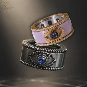 May include: Two wide band rings featuring an evil eye motif. The top ring is light purple with gold edges, a gold eye symbol, and a central blue stone. The bottom ring is dark gray with a black stone eye design and a blue center stone.