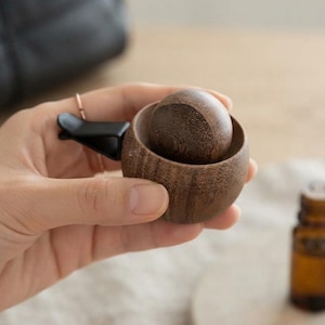 Peut inclure: Un petit diffuseur rond en bois brun foncé doté d'un insert sphérique en bois et d'une attache à pince en plastique noir sur le côté. L'objet présente une texture de grain de bois visible et est conçu pour être fixé sur une surface de ventilation.