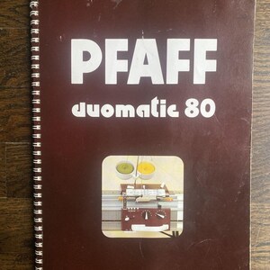 May include: A brown spiral-bound manual with the word "PFAFF" in large white letters and "duomatic 80" below. A rectangular image shows a knitting machine with yellow and green yarn. The cover is slightly worn.