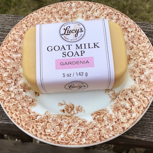 May include: A bar of white goat milk soap with a label that reads "HANDCRAFTED Lucy's GOAT MILK SOAP GARDENIA 5 oz / 142 g".