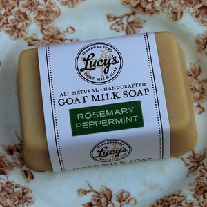 May include: A bar of goat milk soap with a white label that reads "Handcrafted Lucy's Goat Milk Soap Rosemary Peppermint". The soap is on a white plate with a brown floral pattern.