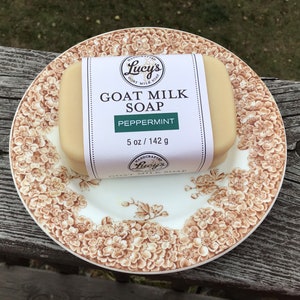 May include: A bar of goat milk soap labeled "Lucy's Goat Milk Soap Peppermint" rests on a floral patterned plate. The soap is 5 oz / 142 g. The plate has a brown and white floral design.