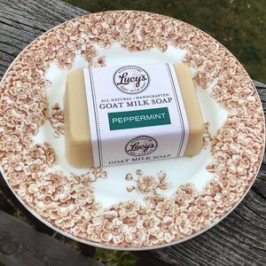 May include: A bar of Lucy's Goat Milk Soap, labeled "Peppermint", rests on a vintage plate with a brown floral pattern. The soap is rectangular, wrapped in a white label with green text. The plate is white with brown floral designs.