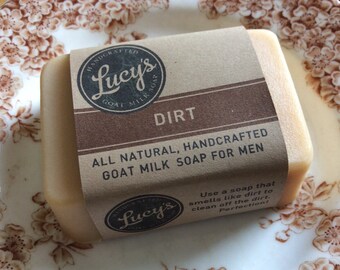 May include: A bar of brown goat milk soap with a brown paper label that reads "HANDCRAFTED Lucy's GOAT MILK SOAP DIRT ALL NATURAL, HANDCRAFTED GOAT MILK SOAP FOR MEN Use a soap that smells like dirt to clean off the dirt. Perfection!"