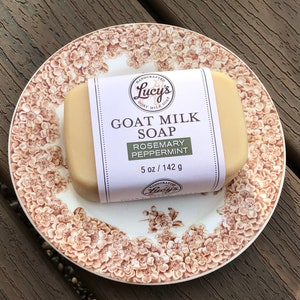 May include: A bar of goat milk soap with a white label that reads "HANDCRAFTED Lucy's GOAT MILK SOAP ROSEMARY PEPPERMINT 5 oz / 142 g". The soap is on a white and brown floral patterned plate.