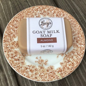 May include: A bar of goat milk soap with a brown label that reads "Handcrafted Lucy's Goat Milk Soap, Almond, 5 oz / 142 g". The soap is sitting on a white plate with a brown floral pattern.