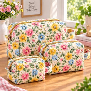 Puede incluir: Un juego de tres neceseres acolchados de distintos tamaños, confeccionados en tela de color crema con un estampado floral repetitivo de flores rosas, amarillas y azules con hojas verdes. Cada neceser tiene una cremallera dorada.