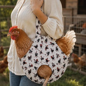 May include: A white fabric carrier bag featuring a repeating pattern of black and red roosters. The bag has a shoulder strap and a circular opening at the bottom, designed to hold a live brown chicken with its head and legs protruding from the bag.