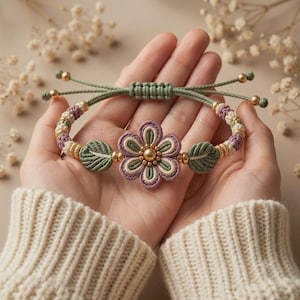 May include: A macrame bracelet featuring a central flower motif with mauve, sage green, and cream cord. The design includes two leaf-shaped accents, small gold-toned metallic beads, and an adjustable sliding knot closure with gold-toned end beads.