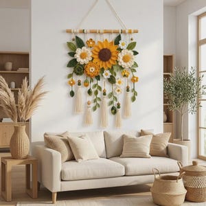 May include: A wall hanging featuring crocheted sunflowers, daisies, and green leaves attached to a horizontal wooden dowel. The piece includes hanging yarn tassels, small wooden beads, and various floral elements in shades of yellow, white, and green.