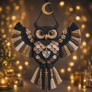 May include: A macrame owl wall hanging featuring black, brown, and cream cord with gold accents. The owl has large amber eyes, a gold beak, and a dangling heart-shaped crystal pendant. A gold crescent moon sits at the top of the hanging cord.