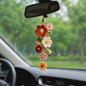 May include: A hanging car accessory featuring a cluster of crocheted flowers in shades of orange, white, red, pink, and yellow, accented with green crocheted leaves and a small gold-toned bell, suspended from a rearview mirror by a thin cord.