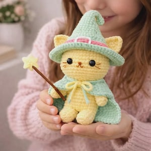 May include: A yellow crocheted cat figurine wearing a light green pointed hat with a pink band and a matching light green cape. The cat has black bead eyes, embroidered whiskers, and holds a brown wand topped with a small yellow crocheted star.