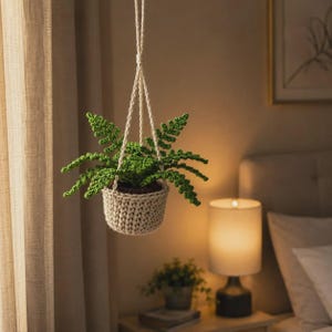 May include: A crocheted hanging plant holder in a light beige color containing a vibrant green crocheted fern. The plant holder features a sturdy woven basket base and long, braided cords that meet at a single point for suspension from above.