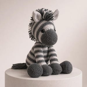 May include: A crocheted zebra stuffed animal featuring alternating horizontal stripes of dark charcoal gray and off-white yarn. The plush toy has a textured mane, two pointed ears, a rounded snout with black button eyes, and a short tail.
