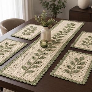 May include: A set of four rectangular crochet placemats and one long matching table runner. Each piece is made of cream-colored yarn with a sage green crocheted leaf vine appliqué in the center and a scalloped sage green border trim.