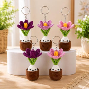 May include: Five crochet keychains featuring small potted flowers with smiling faces. The flowers have green stems and petals in shades of purple, light purple, and pink. Each plant sits in a brown pot with a white base and a metal keyring attached.