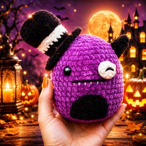 Puede incluir: Un peluche redondo de ganchillo morado con un sombrero de copa negro con banda blanca, un ojo negro, un parche circular blanco sobre el otro ojo, una pequeña boca negra con dientes blancos visibles y un parche negro en la parte inferior.