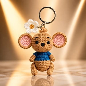 May include: A crochet keychain featuring a tan mouse-like creature with large round ears, pink inner ear details, and a blue shirt. The figure has black eyes, a small black nose, and a white flower with a yellow center attached to the top.