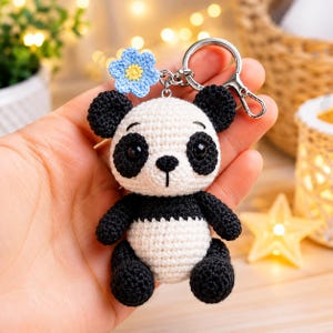Può includere: Un portachiavi a forma di panda all'uncinetto composto da una figura imbottita bianca e nera con orecchie, macchie oculari, braccia e gambe nere. Un piccolo fiore all'uncinetto azzurro con il centro giallo è fissato sulla testa del panda.