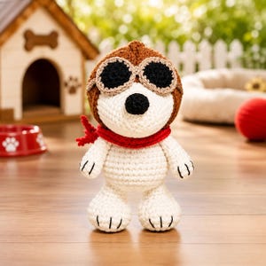 May include: A crocheted stuffed animal resembling a white dog with a black nose, wearing a brown aviator cap with black goggles and a bright red scarf around its neck. The figure is crafted with visible yarn stitches and has black embroidered paw details.