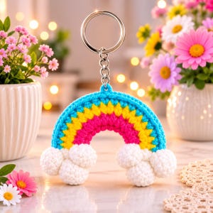 Könnte beinhalten: Ein gehäkelter Regenbogen-Schlüsselanhänger mit einem leuchtend blauen äußeren Bogen, einem gelben Mittelstreifen und einem rosa inneren Streifen. Der Regenbogen endet an beiden Seiten in weißen gehäkelten Wolken und ist an einem silbernen Metallring befestigt.