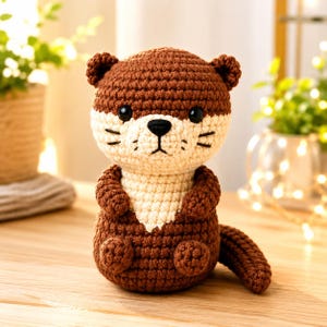 May include: A brown and cream-colored crochet otter plush toy. The otter features a rounded head with small ears, black safety eyes, a black embroidered nose, and stitched whiskers. It has a cream-colored chest patch and a curved tail.