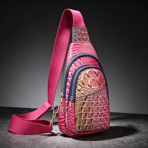Puede incluir: Un bolso bandolera rosa con un patrón de piel de cocodrilo texturizado con degradados metálicos dorados y rosas. El bolso incluye una correa de hombro de tela rosa ancha, ribetes de cremallera azul oscuro y cremalleras de metal dorado.