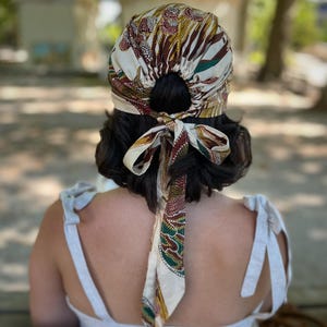 May include: A hair wrap featuring a cream-coloured base with a multi-coloured abstract pattern in shades of brown, green, and yellow. The fabric is gathered at the back with a circular opening and secured with a tied bow featuring two long fabric tails.