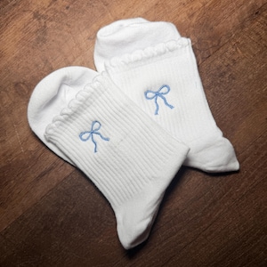 May include: A pair of white ankle socks featuring a ribbed texture and a scalloped lettuce-edge cuff. Each sock is embroidered with a small, light blue bow centered on the upper portion of the ribbed fabric, providing a simple and clean aesthetic.