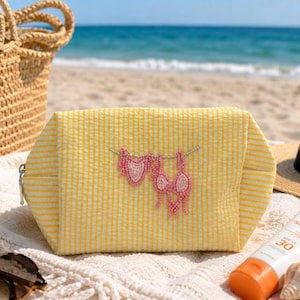 May include: A yellow and white seersucker zippered pouch featuring a beaded design of two pink bikinis hanging on a line. The pouch has a rectangular shape with a silver zipper pull and a textured fabric surface with thin vertical stripes.