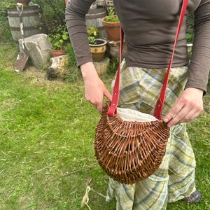 Puede incluir: Un bolso de mimbre marrón tejido con una correa de cuero roja. El bolso tiene forma de media luna y un interior de color claro. La correa está sujeta a la bolsa con remaches.