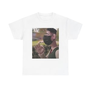 May include: A white t-shirt featuring a centered graphic print of a claymation-style character wearing a black face mask and making an okay hand gesture. The text #FAIRS is printed in black in the upper left corner of the rectangular graphic design.