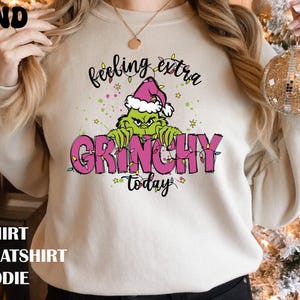 May include: A sand-coloured crewneck sweatshirt featuring a graphic of a green character wearing a pink Santa hat. The text reads "feeling extra GRINCHY today" in black and pink lettering, surrounded by colourful Christmas light illustrations.