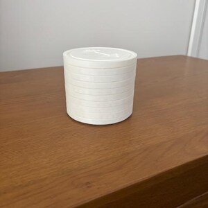 Peut inclure: Un r&eacute;cipient cylindrique blanc au design empilable compos&eacute; de plusieurs couches horizontales. Le couvercle sup&eacute;rieur est plat et circulaire, affichant un petit graphique en relief repr&eacute;sentant une ic&ocirc;ne d'halt&egrave;re au centre de la surface.