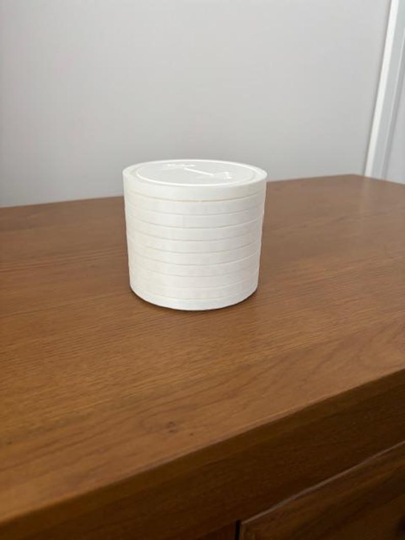 Peut inclure: Un r&eacute;cipient cylindrique blanc au design empilable compos&eacute; de plusieurs couches horizontales. Le couvercle sup&eacute;rieur est plat et circulaire, affichant un petit graphique en relief repr&eacute;sentant une ic&ocirc;ne d'halt&egrave;re au centre de la surface.