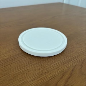 Peut inclure: Un dessous de verre circulaire blanc avec une surface centrale lisse et plate et un rebord ext&eacute;rieur sur&eacute;lev&eacute; et arrondi. L'objet est d'un blanc uni et pr&eacute;sente un design simple et &eacute;pur&eacute; avec un bord distinct qui d&eacute;finit le p&eacute;rim&egrave;tre de la forme.
