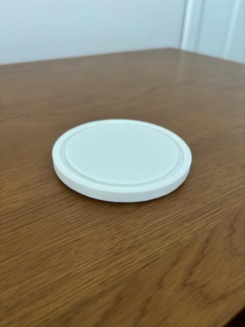 Peut inclure: Un dessous de verre circulaire blanc avec une surface centrale lisse et plate et un rebord ext&eacute;rieur sur&eacute;lev&eacute; et arrondi. L'objet est d'un blanc uni et pr&eacute;sente un design simple et &eacute;pur&eacute; avec un bord distinct qui d&eacute;finit le p&eacute;rim&egrave;tre de la forme.