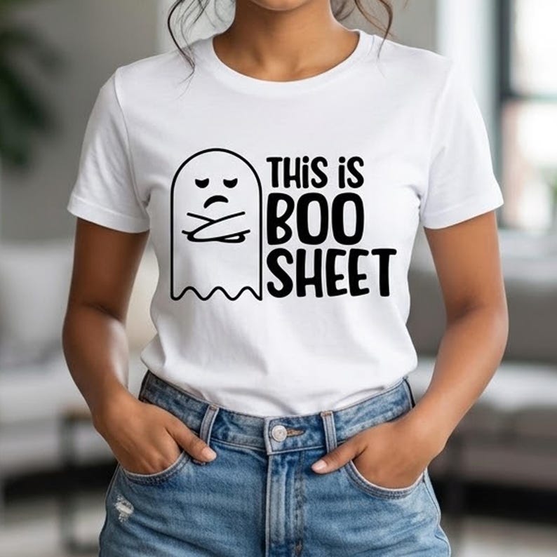 May include: A white short-sleeved t-shirt featuring a black graphic of a grumpy ghost with crossed arms. Next to the ghost, the text reads THIS IS BOO SHEET in bold, black, sans-serif capital letters. The shirt is paired with blue denim jeans.
