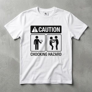 May include: A white short-sleeved t-shirt featuring a black graphic print. The design includes the text CAUTION and CHOCKING HAZARD, along with two pictograms: one showing a person holding a lollipop and another showing a person performing the Heimlich maneuver.