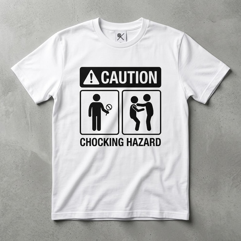 May include: A white short-sleeved t-shirt featuring a black graphic print. The design includes the text CAUTION and CHOCKING HAZARD, along with two pictograms: one showing a person holding a lollipop and another showing a person performing the Heimlich maneuver.