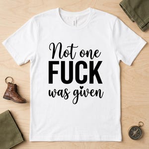 May include: A white short-sleeved t-shirt featuring the black printed text 'Not one FUCK was given' with a small heart icon above the word 'was'. The shirt has a crew neckline and is displayed flat against a light-colored wooden surface.