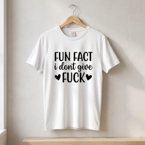May include: A white short-sleeved t-shirt featuring black text that reads FUN FACT i dont give FUCK with a small black heart symbol on each side of the word FUCK. The garment is displayed on a wooden hanger against a neutral wall background.