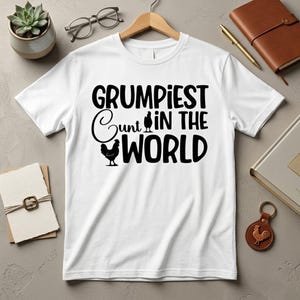 May include: A white short-sleeved t-shirt featuring black text that reads GRUMPIEST CUNT IN THE WORLD. The design includes two black chicken silhouettes, with one replacing the letter U in the word Cunt and another below the word.