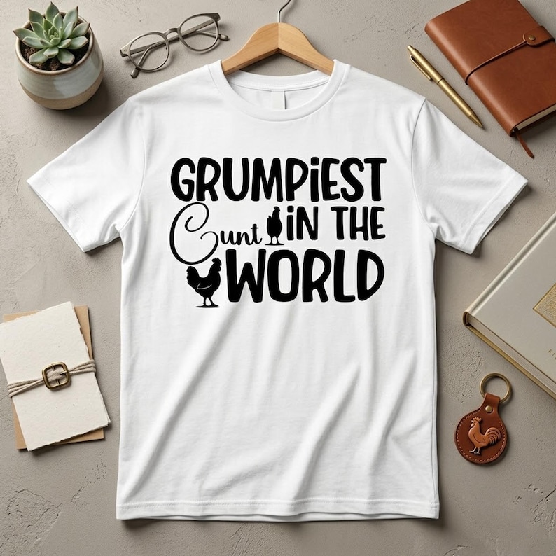 May include: A white short-sleeved t-shirt featuring black text that reads GRUMPIEST CUNT IN THE WORLD. The design includes two black chicken silhouettes, with one replacing the letter U in the word Cunt and another below the word.