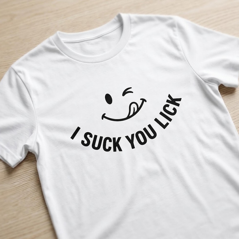 May include: A white short-sleeved t-shirt featuring a black graphic of a winking smiley face with a tongue sticking out. Below the smiley face, the text "I SUCK YOU LICK" is printed in a bold, black, sans-serif font arranged in a slight arc.